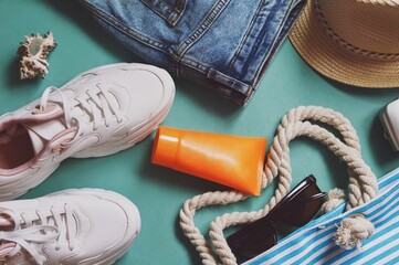 Travel bag essentials for women. White sneakers, blue jeans, straw hat, sunscreen tube, striped cotton bag and sunglasses. Flat lay lifestyle photography. Pack for summer vacation