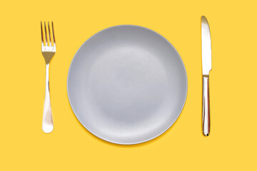 Gray plate and knife with a fork on a yellow background. Empty gray plate and dishes in the form of a knife and fork on an isolated yellow background. Restaurant concept or blank for food placement