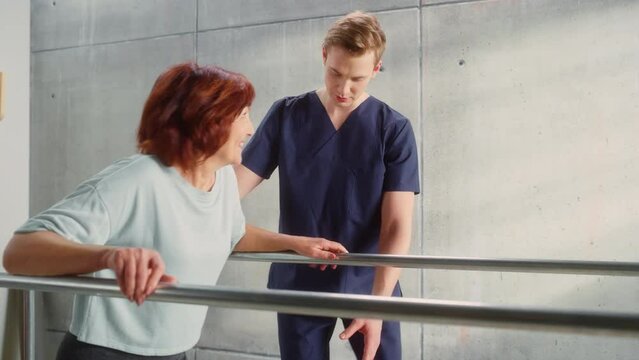 Portrait of Strong Senior Woman with Leg Injury Successfully Walks Holding Parallel Bars. Professional Physiotherapist Assists, Helps, Trains Disabled Person Do Rehabilitative Physiotherapy.