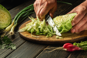 Chef cuts fresh cabbage with knife for salad on a kitchen table with fresh vegetables. Cooking and restaurant or hotel concept