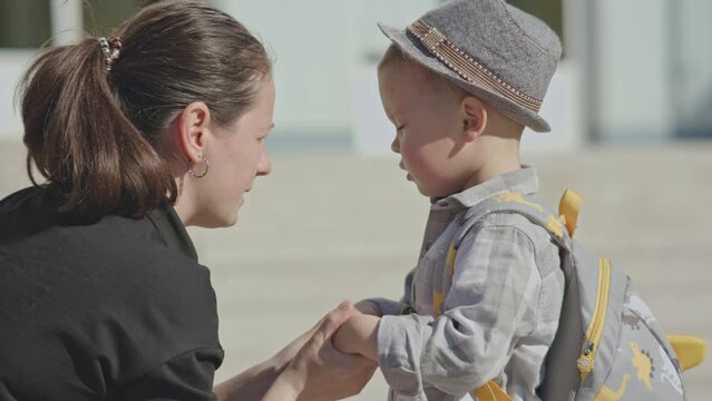 back to school concept, mother sending baby boy child to school first time. caucasian female checking clothes and school bag of preschooler kid, hugs her kid, waving hands, encouraging son in front of