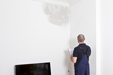 A janitor or custodian inspecting a big water stain and various cracks in wall and ceiling of the...