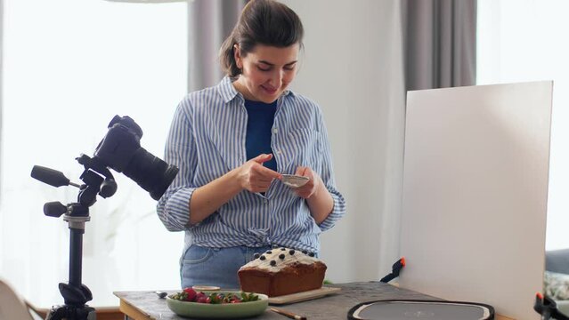 blogging, cooking and people concept - happy smiling female photographer or food blogger with camera pouring powdered sugar to cake in kitchen at home