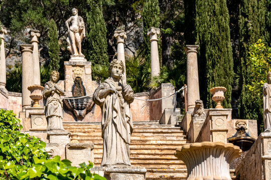 Horizontal photography of a female sculpture in front of an old staircase with sculptures and amphorae called Apollo Staircase with the statue of god Apollo in the background in the public Raixa manor