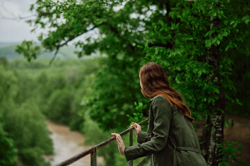 Naklejka na ściany i meble red-haired woman walking in the park looks at the stormy mountain river. Travel, rainy weather