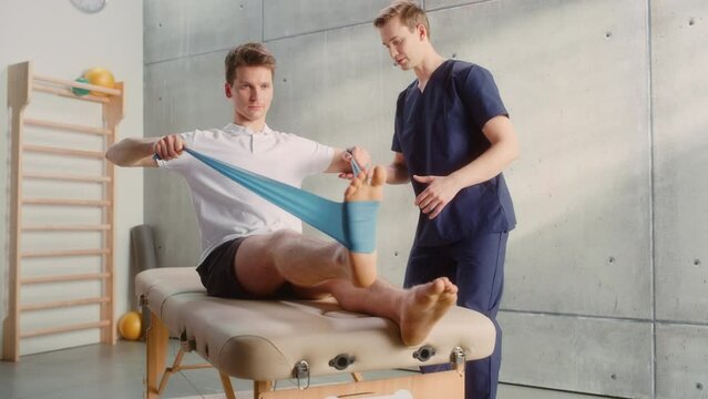 Young Male Athlete Undergoing Physiotherapy, Professional Sport Masseur Helping with Foot Exercise with a Rubber Band. Musculoskeletal Pain Therapy and Rehabilitation Concept.