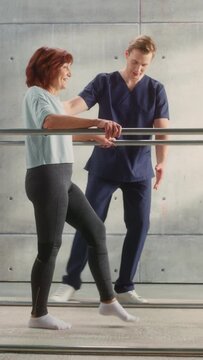 Vertical Portrait of Strong Senior Middle Aged with Leg Injury Successfully Walks Holding Parallel Bars. Professional Physiotherapist Assists, Trains Disabled Person Do Rehabilitative Physiotherapy.