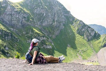 Young woman hiker with purple hair in cap with backpack sitting and looking at the mountain view in summer healthy active lifestyle, outdoor activities, landscape