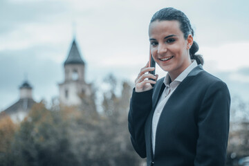 Mujer empresaria de negocios formal con seriedad y de pie y llamando por teléfono móvil y hablando con una sonrisa en el centro de la ciudad parada por la calle para llegar a su trabajo - obrazy, fototapety, plakaty