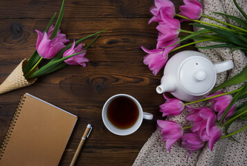 Tulips, a notebook, a white teapot and a cup of tea are laid out on a wooden table