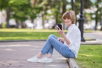 happy elderly woman in white shirt enjoying music on mobile phone through headphones in city park. woman 60 years old listens to music, and rests in the park outdoors