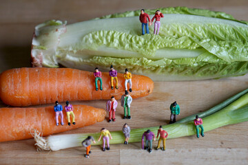 Miniature figurine character: people sitting on carrots, and spring onions, and some standing on lettuce and wooden floor, concept of fresh organic vegetables for good health in the market 