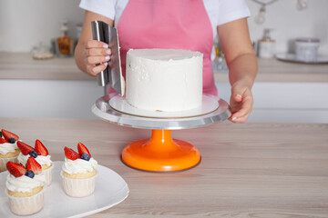 Pastry chef confectioner young caucasian woman in the kitchen. She is decorating the cake on a stand. Nearby are cakes