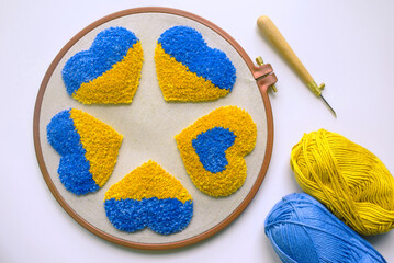 Top view of five tufted hearts on grey fabric in a hoop, punch needle and two balls of yarn in blue...
