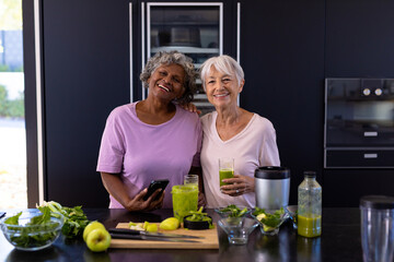 Portrait of smiling multiracial senior female friends with smoothie on counter standing in kitchen