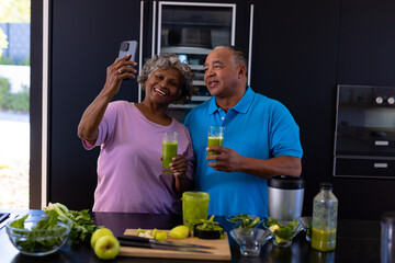 African american senior woman taking selfie with caucasian man while having smoothie in kitchen