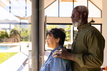 Side view of multiracial senior couple looking through window while standing in retirement home