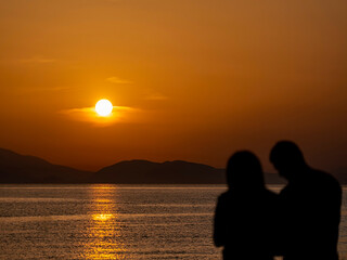 Fototapeta na wymiar A couple at a sea beach with colorful sunset sky. Panoramic beach landscape. Tropical beach and seascape and a distant island in the background. Calmness, tranquil, relaxing sunlight, summer mood.