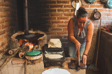 Mujer mexicana torteando maza de maíz en un metate y una estufa de leña para hacer tortillas...