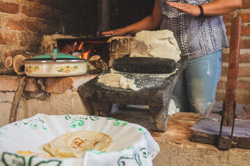 Mujer mexicana torteando maza de maíz en un metate y una estufa de leña para hacer tortillas...