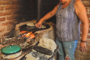 Mujer mexicana torteando maza de maíz en un metate y una estufa de leña para hacer tortillas...