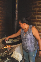 Mujer mexicana torteando maza de maíz en un metate y una estufa de leña para hacer tortillas...