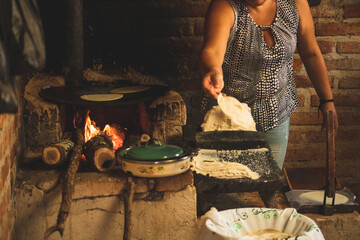 Mujer mexicana torteando maza de maíz en un metate y una estufa de leña para hacer tortillas...