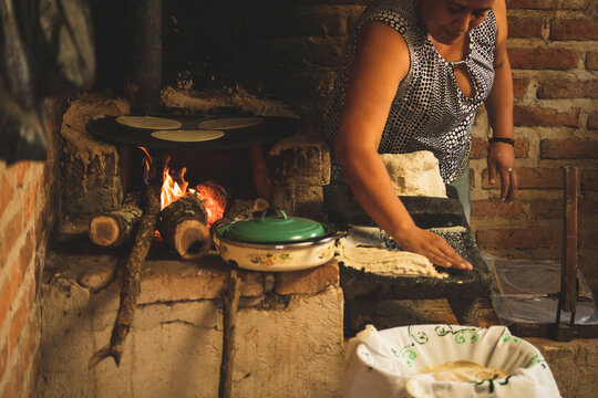 Mujer mexicana torteando maza de maíz en un metate y una estufa de leña para hacer tortillas caseras	