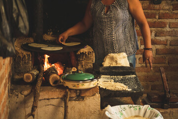 Mujer mexicana torteando maza de maíz en un metate y una estufa de leña para hacer tortillas...