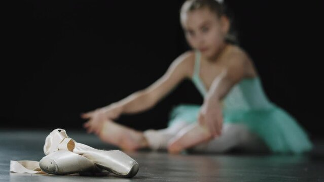 Teenage ballerina girl in blue tutu sits on floor stretching, ballet shoes pointe shoes in foreground. Child learning dancing stretch legs resting after rehearsal dance preparing to perform on stage