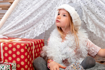 portrait of a cute little girl child wearing a silver color Christmas hat sitting between gift boxes