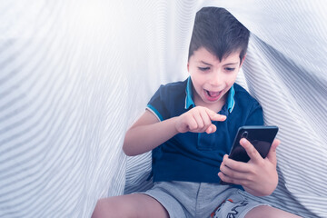 smartphone addiction of a child under white striped sheets holding a smartphone in his hand with very excited expression