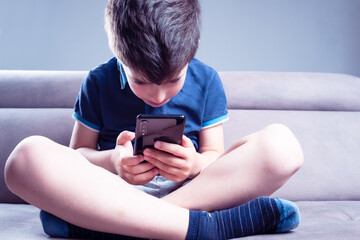 smartphone addiction of a child sitting cross-legged on the tan sofa and looking at the smartphone completely enraptured