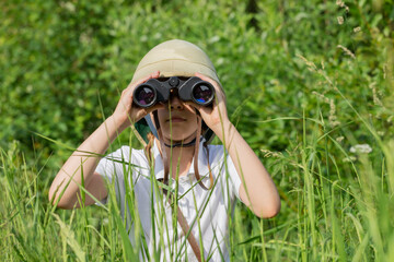 Preteen girl wearing pith helmet hiding in the grass looking through binoculars observing summer...