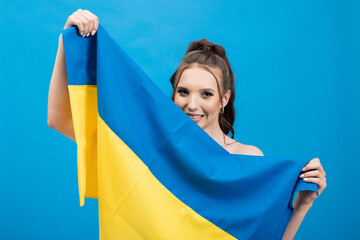 Woman holding yellow and blue flag of Ukraine on studio background. Smiling brunette with hair tied...