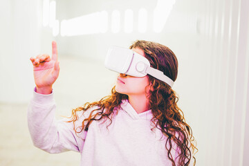 Curly haired young woman in virtual reality glasses moves hands touching online object and looks around standing in white futurisctic tunnel