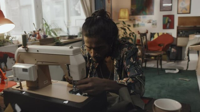 Young male Indian dressmaker with long dark hair in bun working on sewing machine in messy retry style atelier