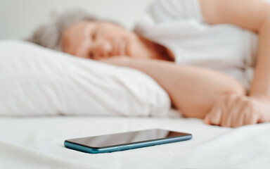 Close-up of mobile phone on bed and sleeping mature woman in bedroom in morning. Selective focus on smartphone