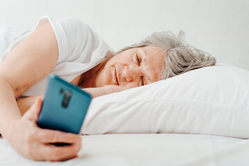 Obraz na płótnie Canvas Mature awakened woman holding mobile phone and looking at screen while lying on bed in bedroom in morning. Elderly woman using smartphone after sleeping indoors, close-up