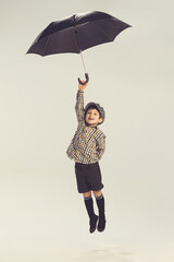 Portrait of little boy, child in checkered shirt and shorts playing, jumping with umbrella isolated over grey studio background © Lustre Art Group 