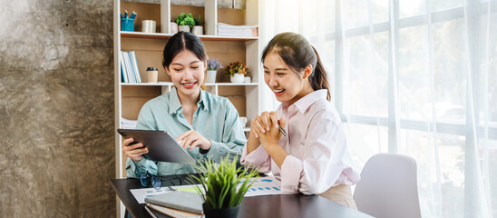 Asian female workmates using digital tablet and documents to look at sales analysis, working on...