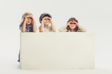 Portrait of little children, boy and two girls peeking out the table isolated over grey studio background