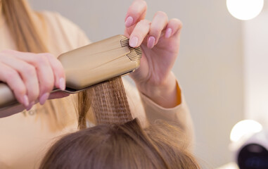 Close-up female hands with a curling iron straightens a strand of hair. The hairdresser makes a hairstyle for a young woman. Barber shop, business concept. Beauty salon, hair care.