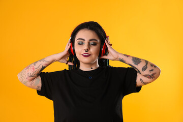 Fototapeta na wymiar Beautiful young woman with tattoos on arms, nose piercing and dreadlocks listening music against yellow background