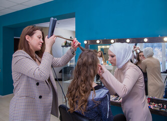 Two masters with a smile curl their hair and make up a girl in the salon. Make-up artist and hairdresser create an image of a young woman. Business concept - beauty salon, facial skin and hair care.