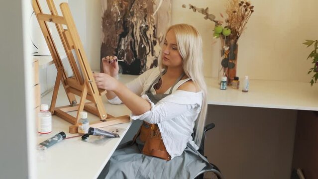 Beautiful girl blonde artist paints an easel while sitting in a wheelchair