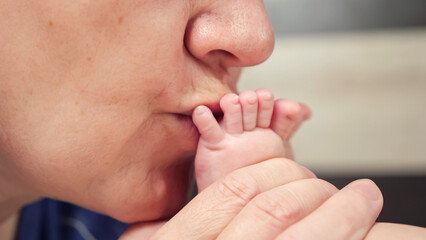 Grandma kisses bare tiny feet with little fingers of newborn baby on blurred background. Woman...