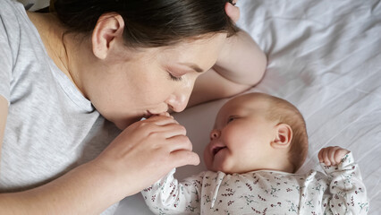 Happy mother talks to newborn daughter holding tiny hand in bedroom. Woman and adorable baby girl lie on white sheet laid on bed at hom, top view