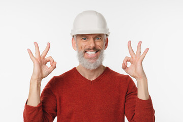 Smiling caucasian mature man builder architect engineer construction worker in white hardhat showing okay gestures looking at camera isolated in white background