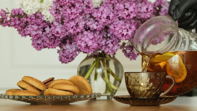 Tea being poured into tea cup. Unrecognizable person pouring hot black tea in cup in morning, vase with a lilac flowers near on table. Breakfast concept. White background, Close up, slow motion